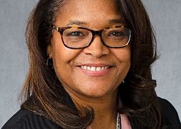 A headshot of Karen Wright. Karen is wearing a black sweater over a light pink shirt, sitting against a gray background